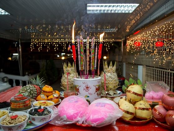 Decorated altar with lit incense sticks and candles, surrounded by offerings of fruits, pastries and food bowls, with sparkling lights in the background during a traditional ceremony.