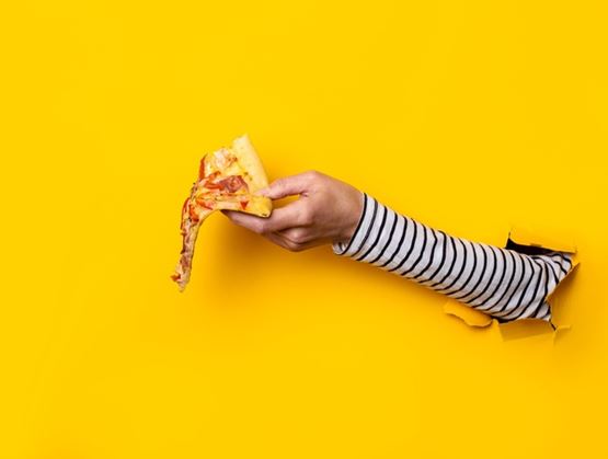 A hand emerging from a torn yellow background holds a slice of pizza with melted, stretchy cheese.