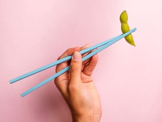 Hand holding blue chopsticks picking up an edamame pod against a pink background.