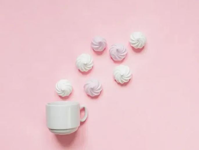 White cup accompanied by small meringues arranged on a pastel pink background, creating a minimalist composition around sweet treats.