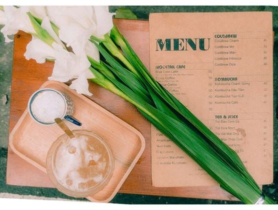 Top view of a table featuring a café menu, an iced drink and a cup, accompanied by white flowers and long green leaves arranged decoratively.