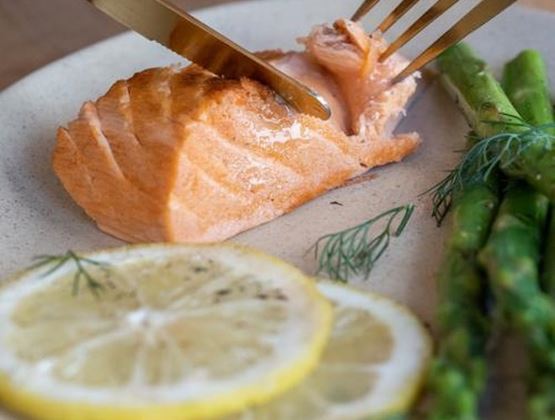 This image shows a piece of salmon being cut with a fork and knife, served with lemon slices and asparagus.