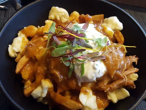 Poutine served in a black bowl, topped with cheese curds, brown gravy, pulled meat, cream, and microgreens.
