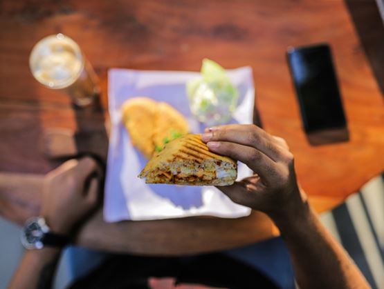 Person holding a grilled sandwich from a top view, with a drink and a smartphone on a wooden table, suggesting a casual dining moment.
