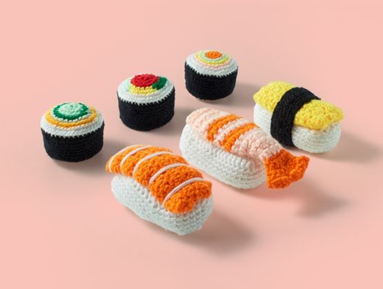 Assortment of sushi and maki made from crochet, arranged on a pink background, creating a playful and handcrafted composition.