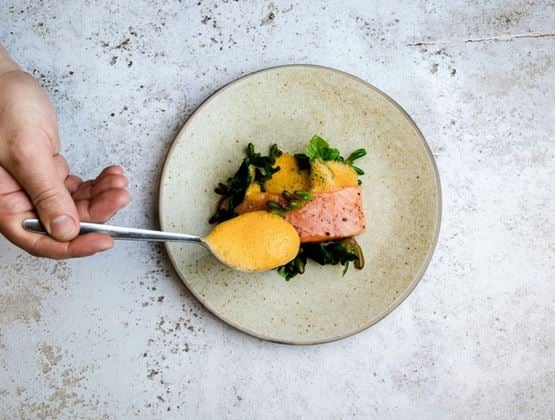 Hand holding a spoonful of orange sauce over a plate of salmon with green vegetables, on a light stone surface.