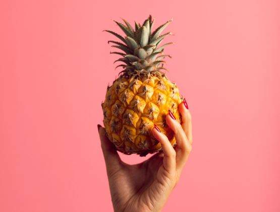 Main tenant un ananas sur fond rose vif, avec ongles rouges.