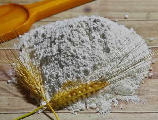 Pile of flour with wheat stalks and a wooden spoon on a rustic wooden surface.