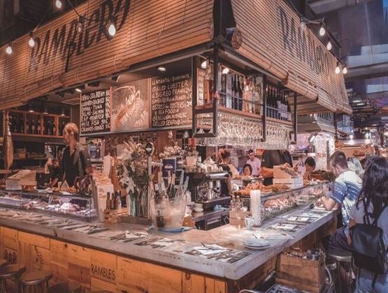 eafood and tapas counter in a bustling food market, with customers seated at the bar and staff preparing dishes behind displays of fresh produce