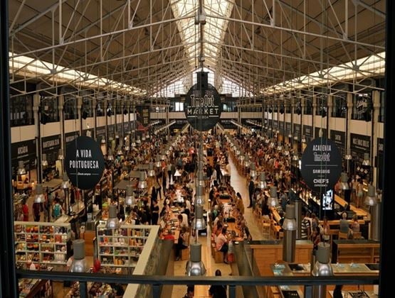 Aerial view of Time Out Market Lisbon, with long communal dining tables crowded with visitors under a steel-framed glass roof, surrounded by food counters
