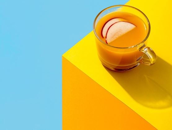 Boisson orangée servie dans une tasse en verre avec des tranches de pomme sur un fond bleu et jaune.