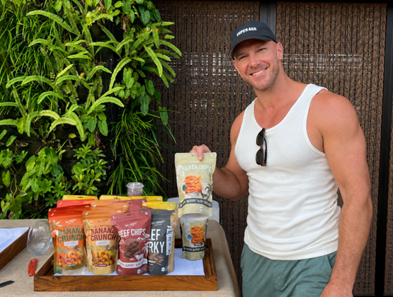 Smiling man presenting Bali Forages snack products beside a display of Banana Crunch, Beef Chips, Beef Jerky and Chicken Chips.