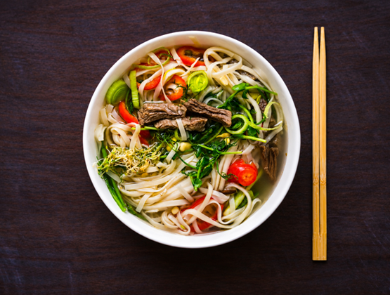 Bowl of noodle soup with beef, vegetables and herbs, served with chopsticks.