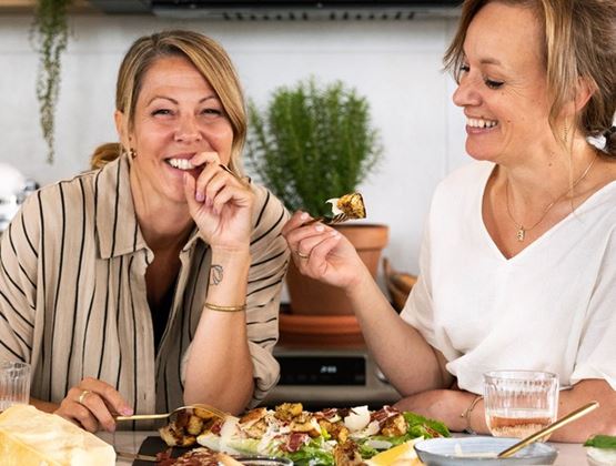 Two women laughing as they share a Caesar salad at a kitchen counter, surrounded by fresh herbs, Parmesan and black garlic.