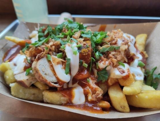 Fries served in a cardboard tray, topped with sauce, cream, fresh herbs, crispy onions, and pieces of sausage.