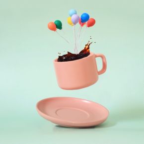 Pink cup floating above a saucer, filled with coffee and colorful balloons rising from it, on a light green background.
