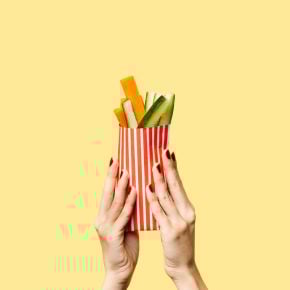 Hands holding a red-and-white striped cone filled with vegetable sticks against a yellow background.