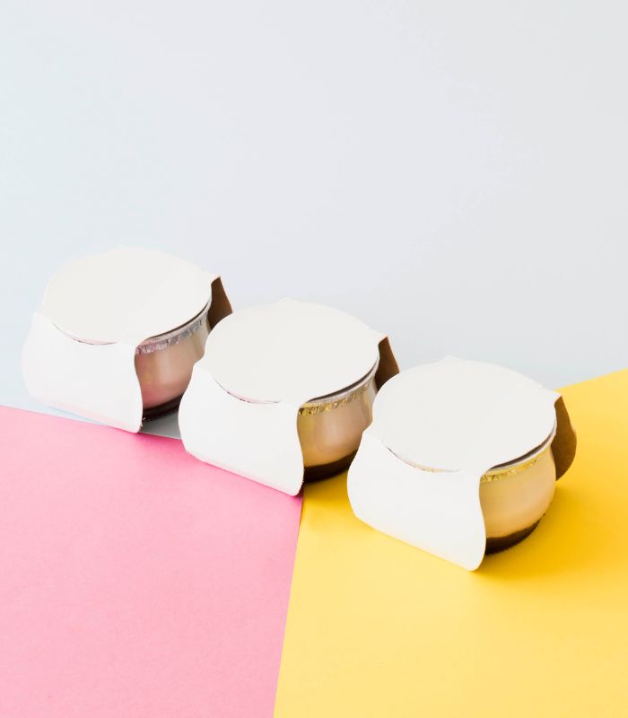Three glass yogurt jars with white lids, packaged in a cardboard holder on a pink, yellow, and white background.