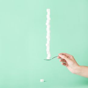 Hand holding a spoon balancing a tall stack of sugar cubes against a mint green background.