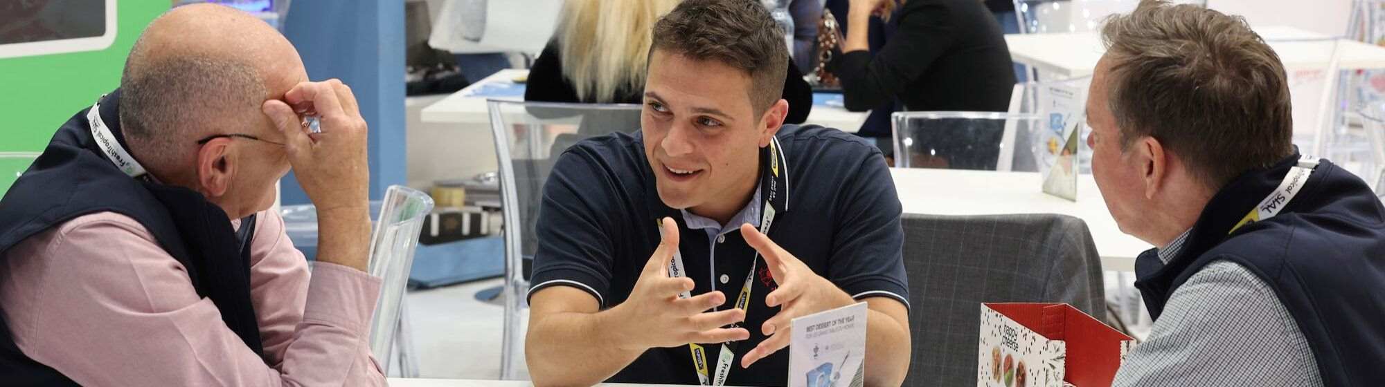 Three men engaged in discussion around a table at a trade show.