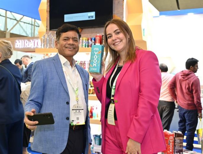 A man and a woman posing at a trade show booth, smiling while showcasing a beverage product. The woman is wearing a pink suit and holding a blue drink container labeled "Yook."