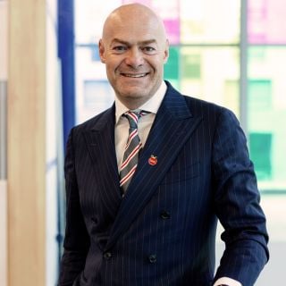 Portrait of a smiling man in a blue pinstripe suit, standing before colorful windows in a bright indoor space.