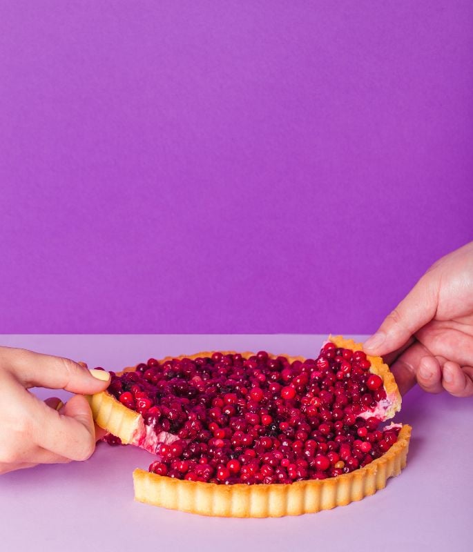 Two hands pulling apart a red berry tart on a minimal purple background