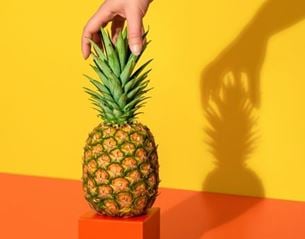 Hand holding a pineapple on yellow and orange background, minimalist composition with sharp shadow and vivid colors