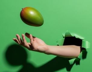 Hand emerging from green background tossing a mango in the air, minimalist scene with contrast between fruit and colored backdrop