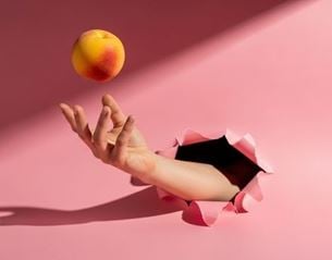 Hand emerging from torn pink background tossing a nectarine in the air, minimalist composition with soft contrast and cast shadow