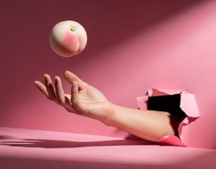 Hand emerging from torn pink background tossing a light peach in the air, minimalist composition with soft light and shadow