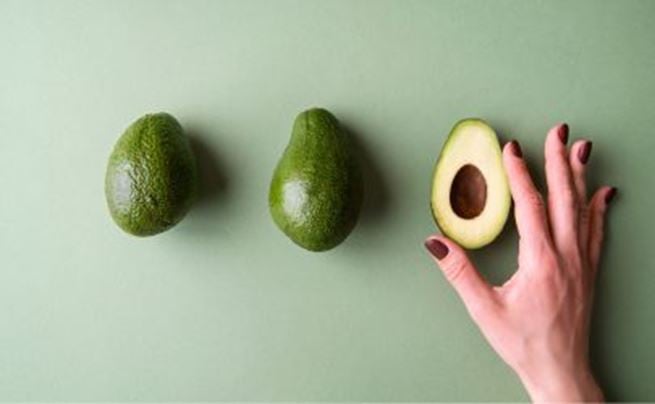 Whole avocados and a halved avocado held by a hand on a green background, minimalist composition.
