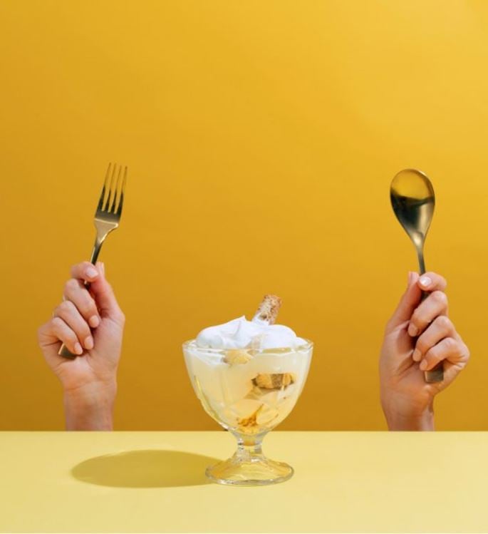 Creamy dessert in a glass with hands holding spoon and fork on a yellow background, indulgent minimal food concept
