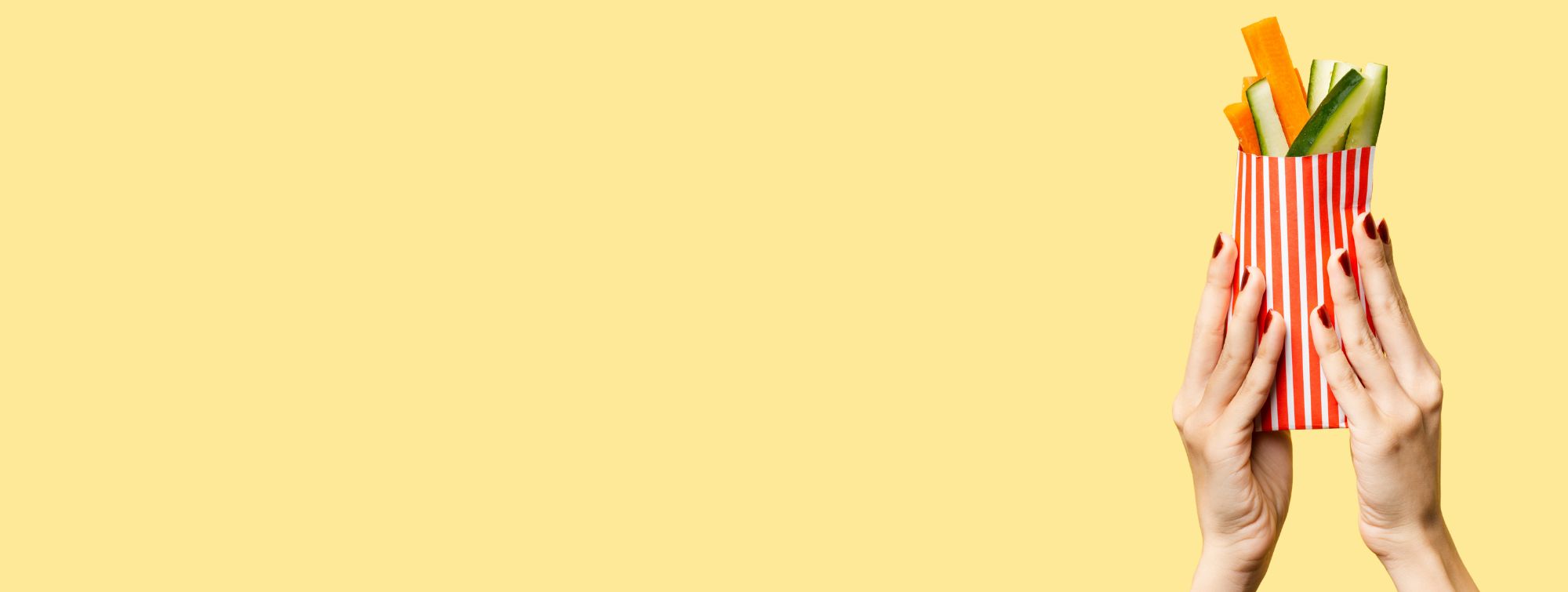 Hands holding a striped container with fresh vegetable sticks on a yellow background with copy space, healthy food concept