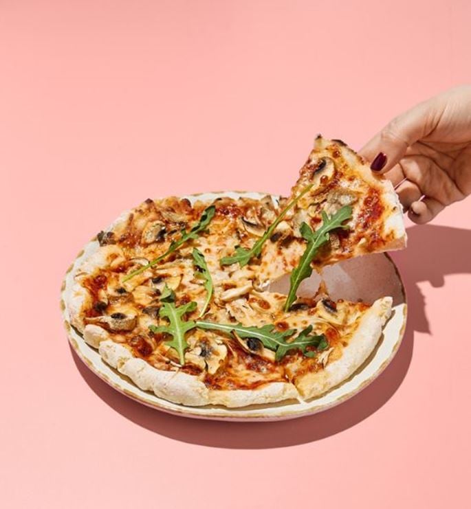 Hand picking up a pizza slice with arugula and mushrooms on pink background, studio shot