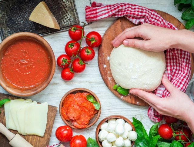 Preparing pizza dough with fresh ingredients, tomatoes, mozzarella and sauce on wooden countertop