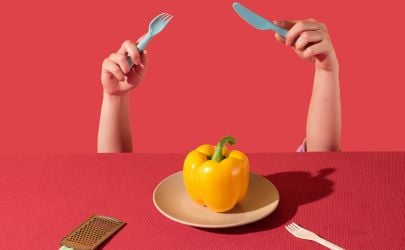 Two hands holding a fork and knife in front of a yellow bell pepper on red background.
