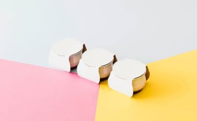 Three small white cream pots aligned on a pastel pink and yellow background.