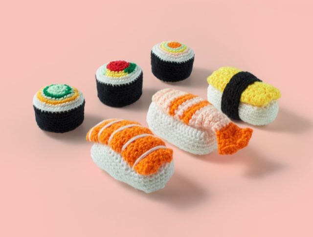 Various crochet yarn sushi pieces arranged on a pastel pink background.