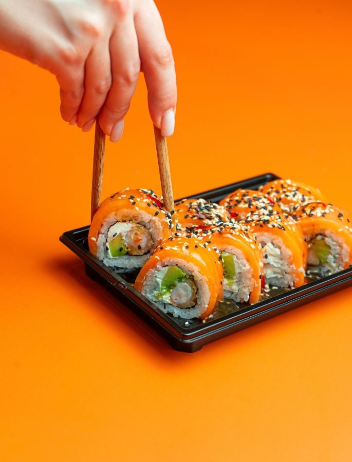 Hand using chopsticks to pick up a salmon sushi from a black tray.