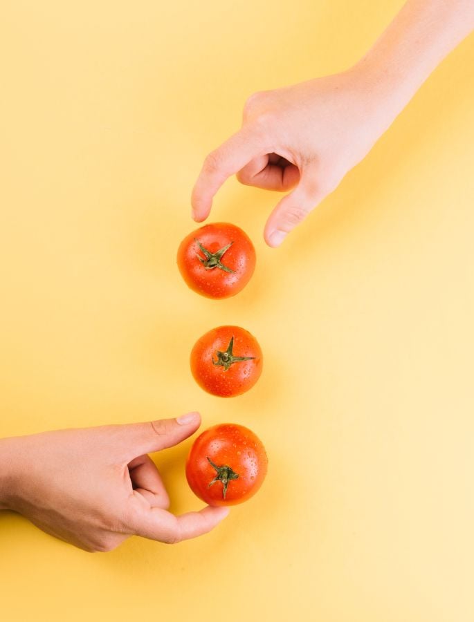 Three red tomatoes vertically aligned between two hands on a yellow background.