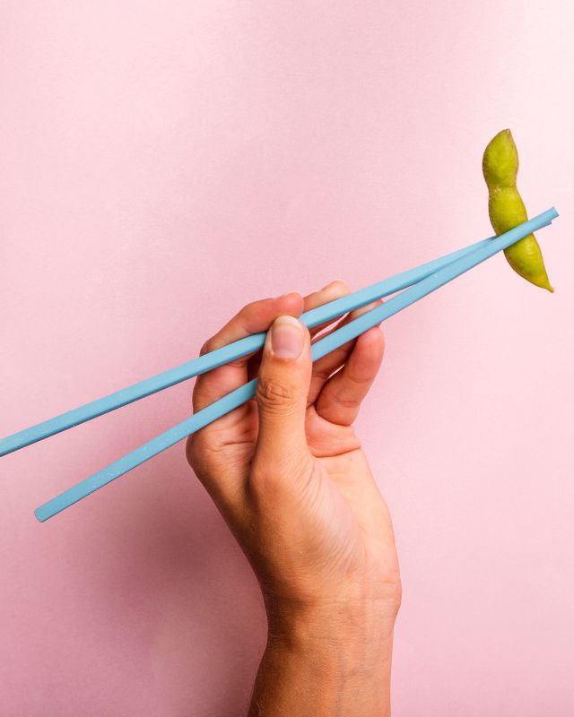 Edamame held with blue chopsticks on pink background