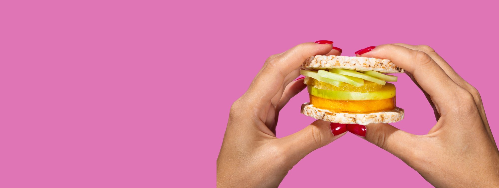 Hands holding a rice cake sandwich filled with fruit slices against a pink background.
