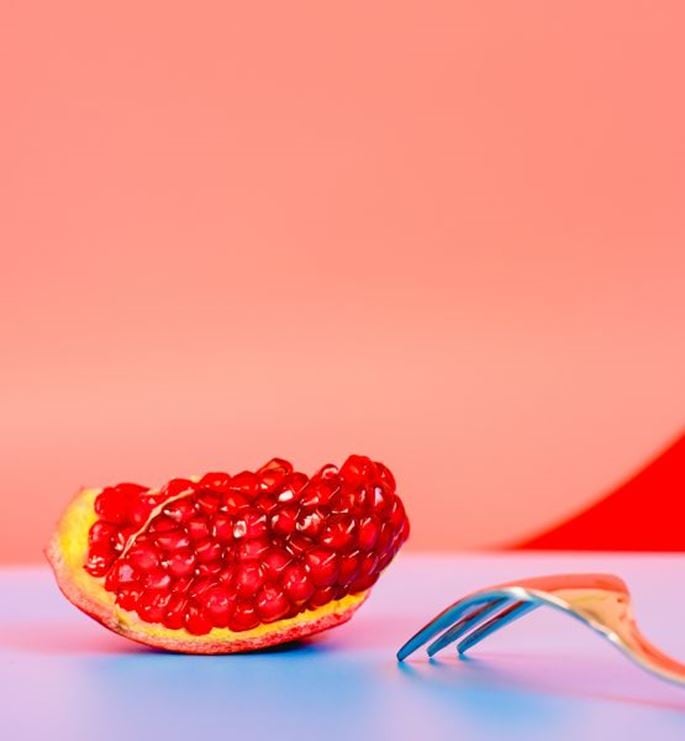 Pomegranate wedge beside a fork against a coral pink background.