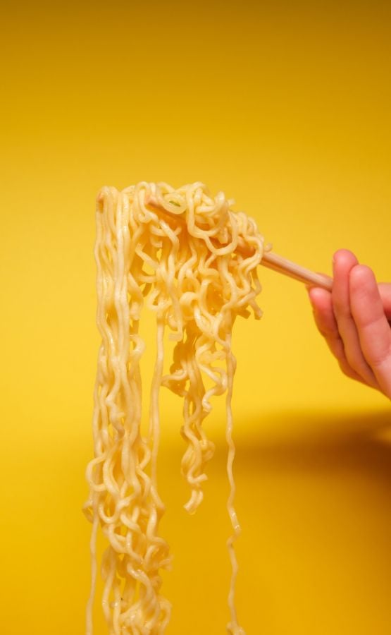 Hand lifting noodles with chopsticks against a yellow background.